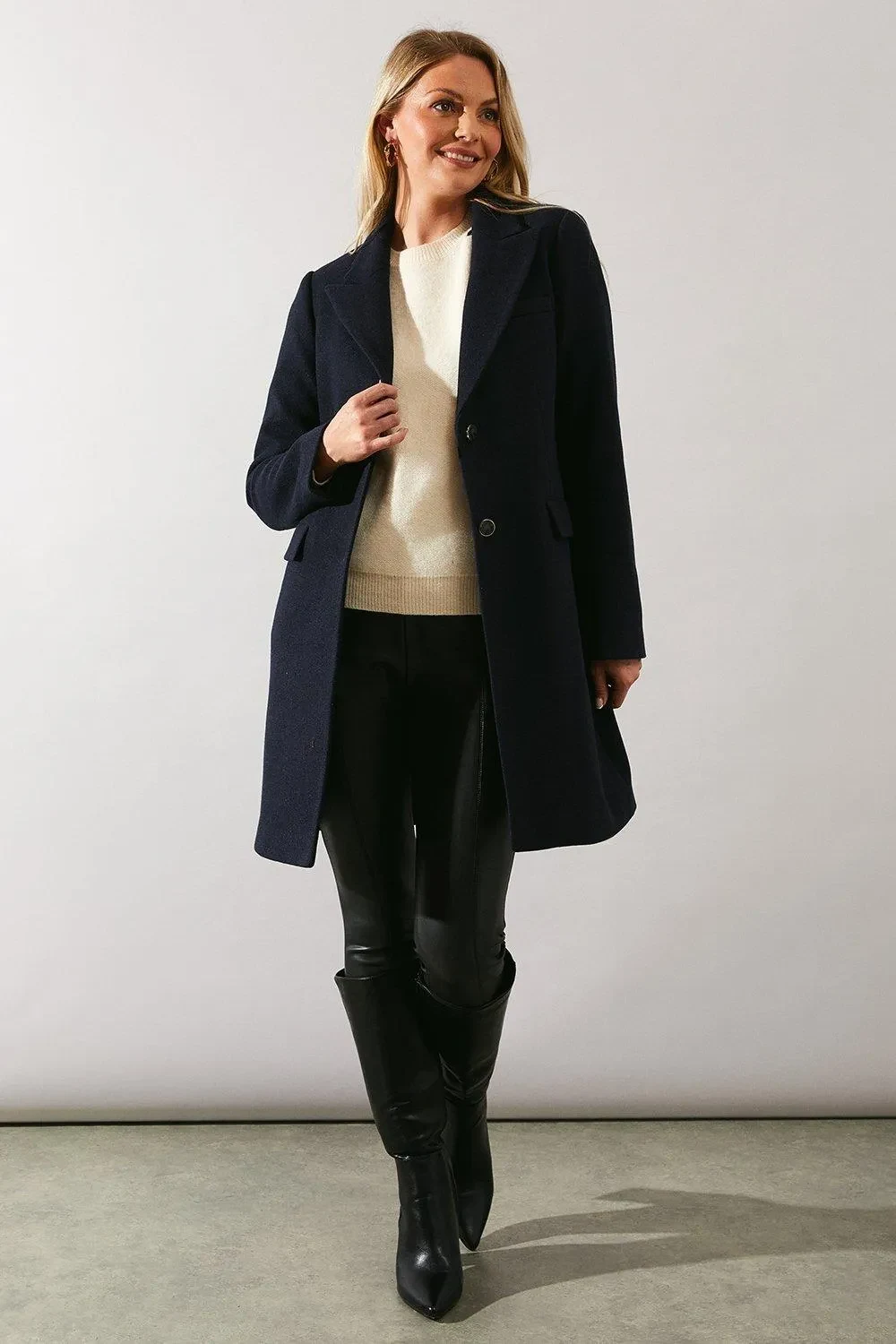 Navy Knit Sweater under a Cream Coat with Black Leather Trousers and Black Knee-high Boots